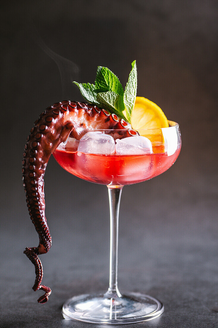 Alcoholic cocktail with ice cubes served with octopus tentacle and lemon slice placed on table against gray background in studio