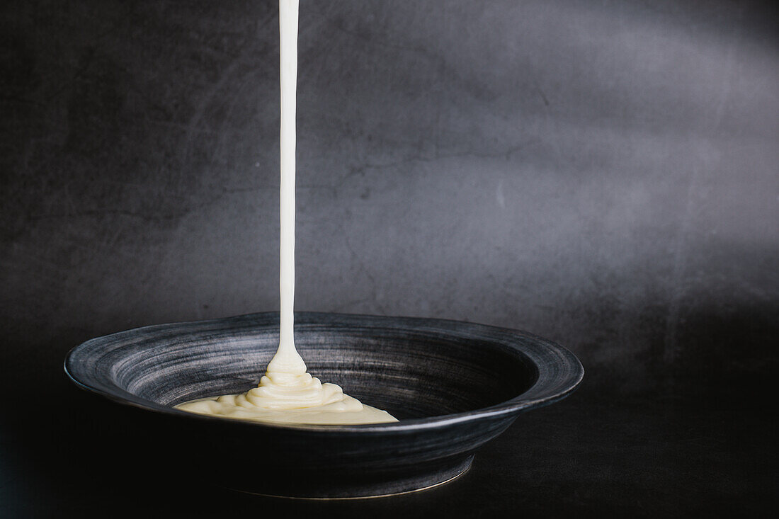 White tasty sauce pouring into dark ceramic bowl placed on table against gray background in light room during cooking process