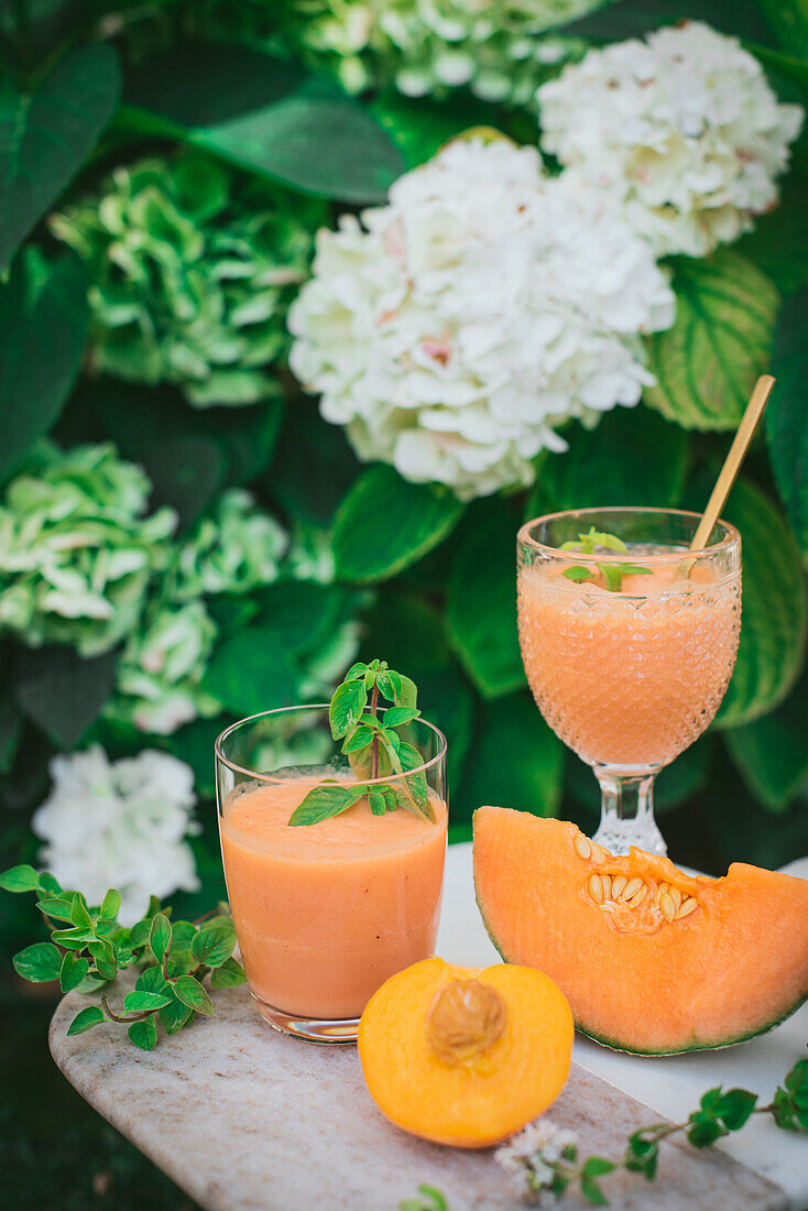 Rockmelon sweet melon and peach slice near smoothie on glass placed on white table in light kitchen on blurred background