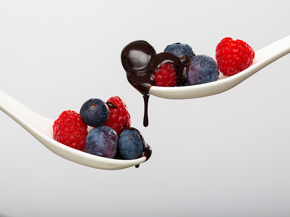 Sweet chocolate topping pouring on heap of ripe fresh raspberries and blueberries on spoons against white background in light studio