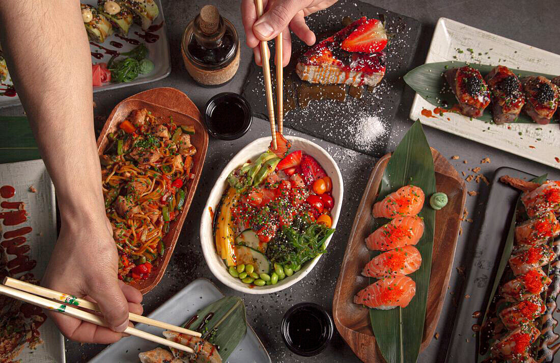 Verschiedene asiatische Gerichte (Sushi, Poke Bowl, Udonnudeln, Jiaozi), Hände mit Essstäbchen