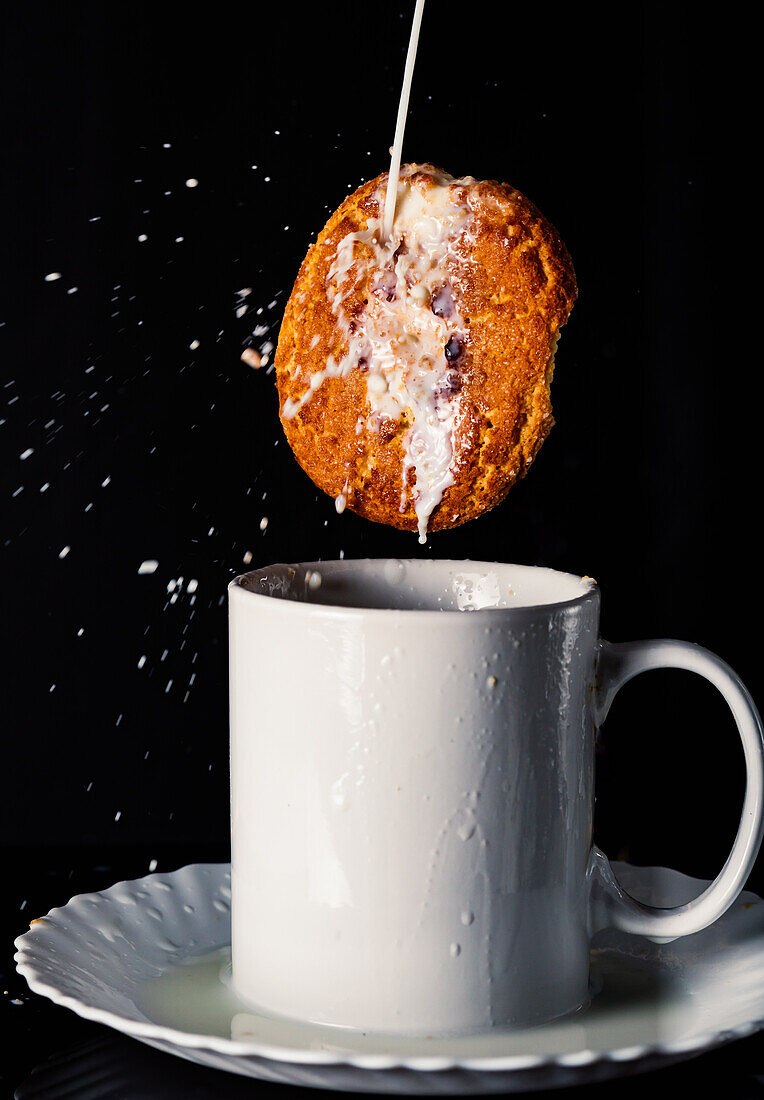 Oatmeal cookie with milk falling in ceramic mug among splatters on black background