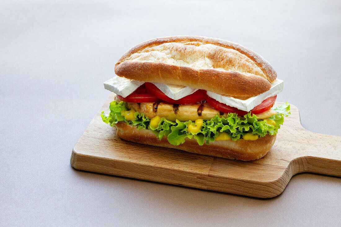 Tasty sandwich with chicken on lettuce and tomato slices with cheese served on wooden chopping board on gray background in kitchen