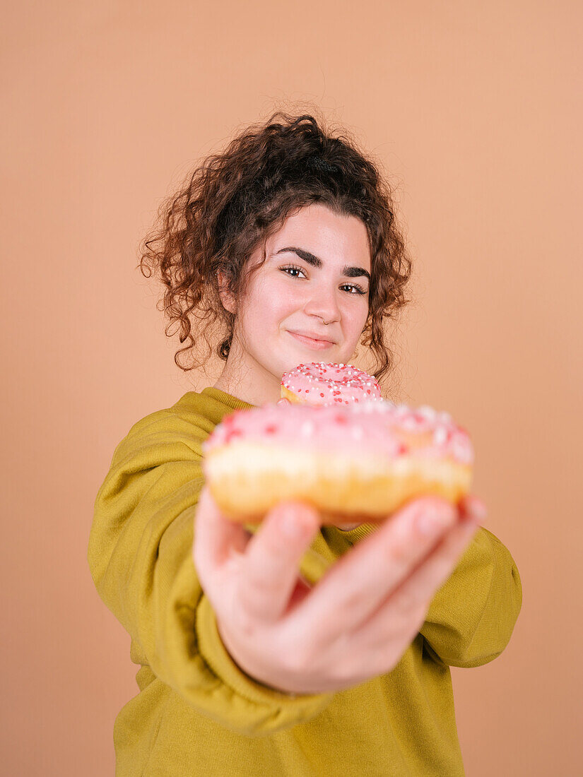 Cheerful young female eating appetizing sweet doughnut and giving to camera in studio against beige background