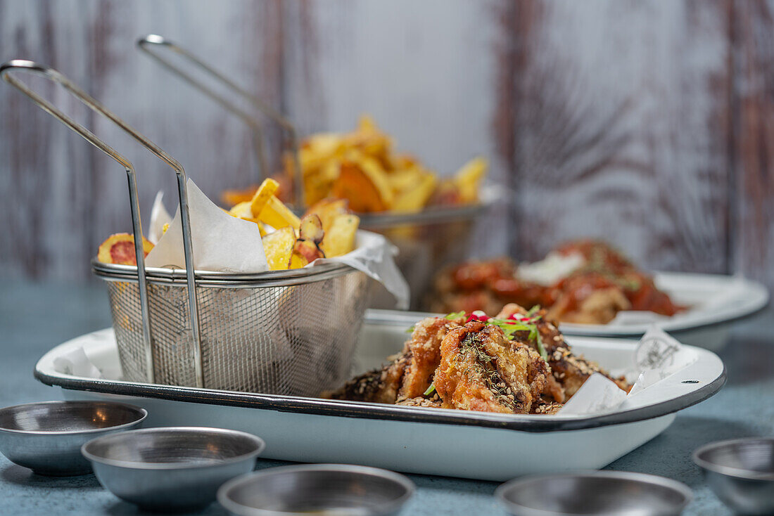 Tasty fried potatoes with breaded chicken wings placed on table with set of sauces in light kitchen