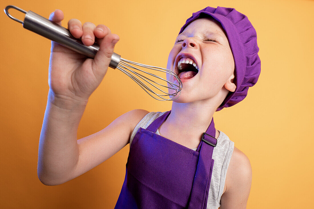 Cheerful boy in purple chef uniform singing into metal whisk with closed eyes while standing against yellow background in light modern studio