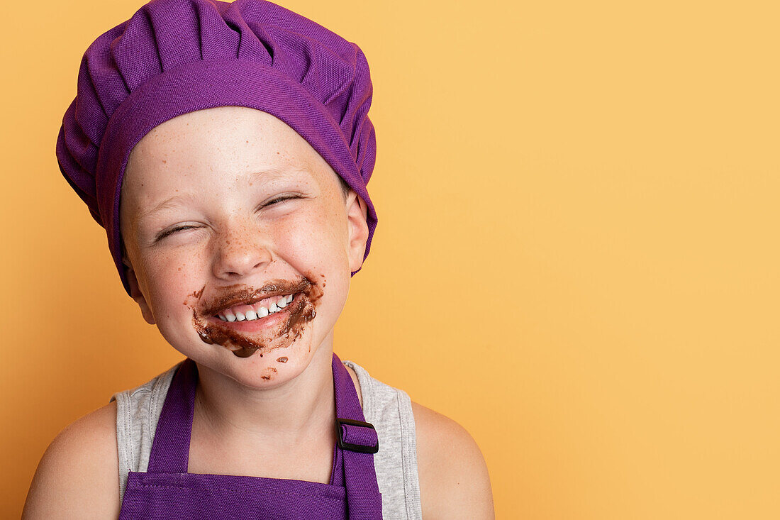Delighted dirty boy in purple chef hat and apron with face covered with chocolate standing against yellow background in studio