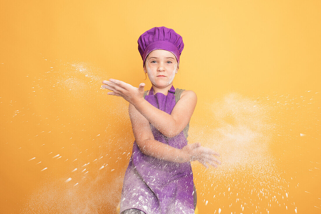 Optimistic girl in purple chef uniform clapping hands with white flour while sitting against yellow background in modern light studio looking at camera