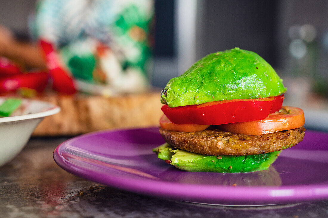 Soft focus of halves of avocado forming burger with vegetables and vegan patty on plate near woman in kitchen at home