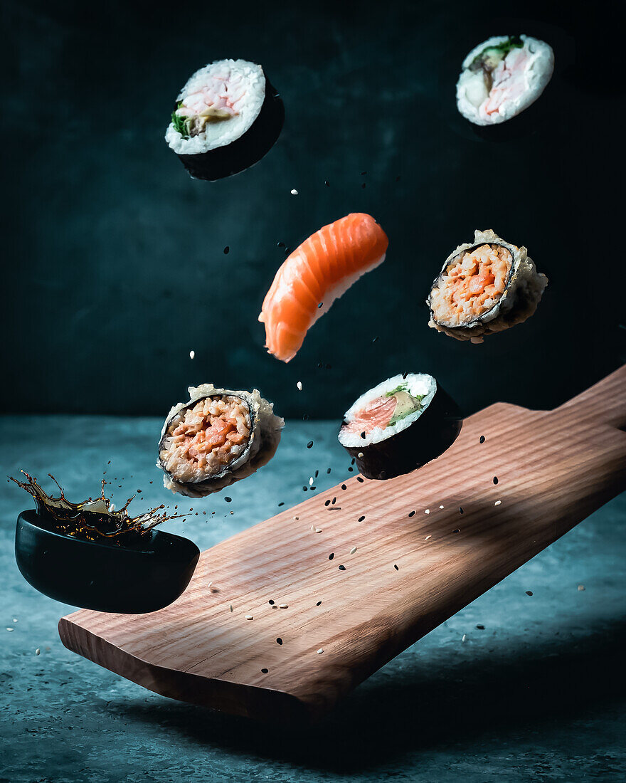 Assorted sushi and a small bowl of soy sauce, flying over wooden board