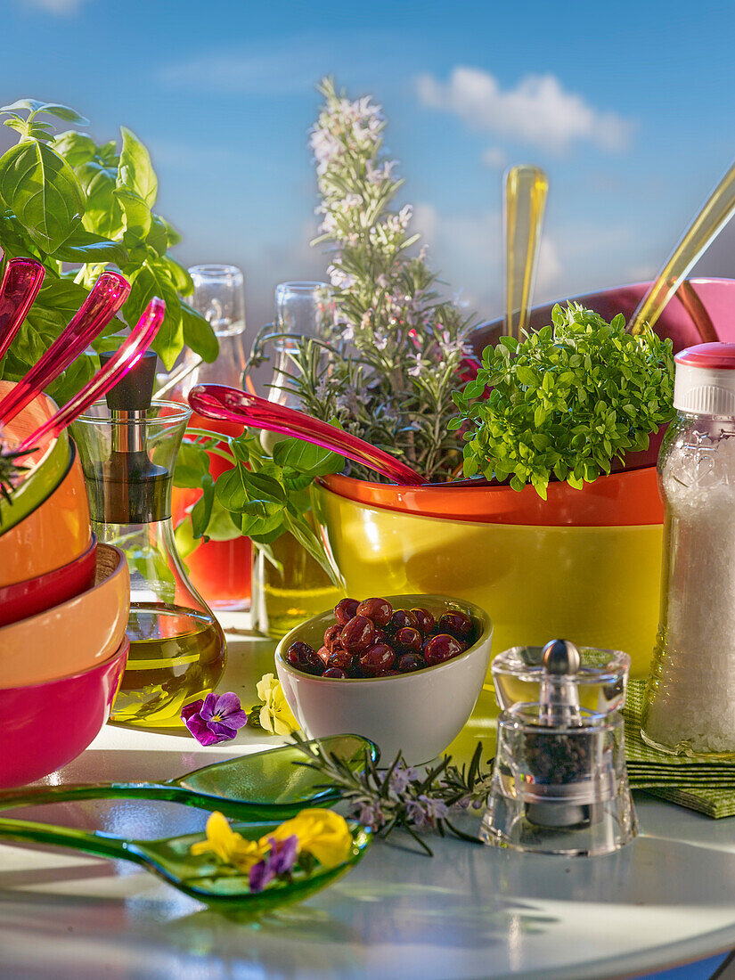 Stillleben mit bunten Salatschüsseln, Salatbesteck, Kräutern, Oliven, Pfeffermühle und Olivenöl
