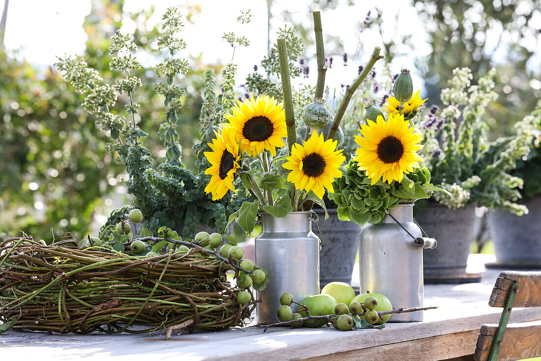 Sunflowers with poppy pods and hydrangea in milk jugs, next to them a wreath of vines