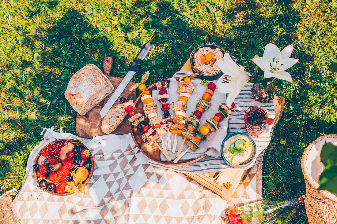 Picknick im Garten: vegetarische Sandwich-Spieße, Brot, Dips und Schale mit frischen Sommerfrüchten