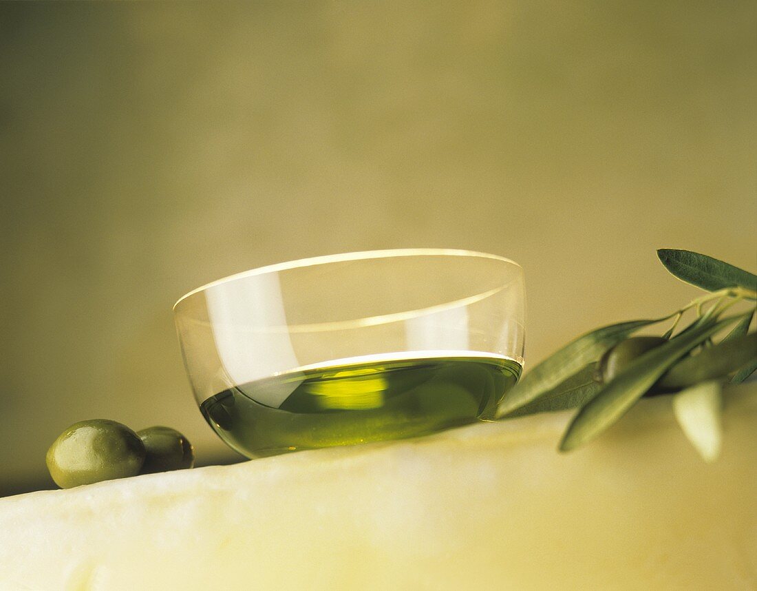 Olive oil in glass bowl, two green olives & olive branch