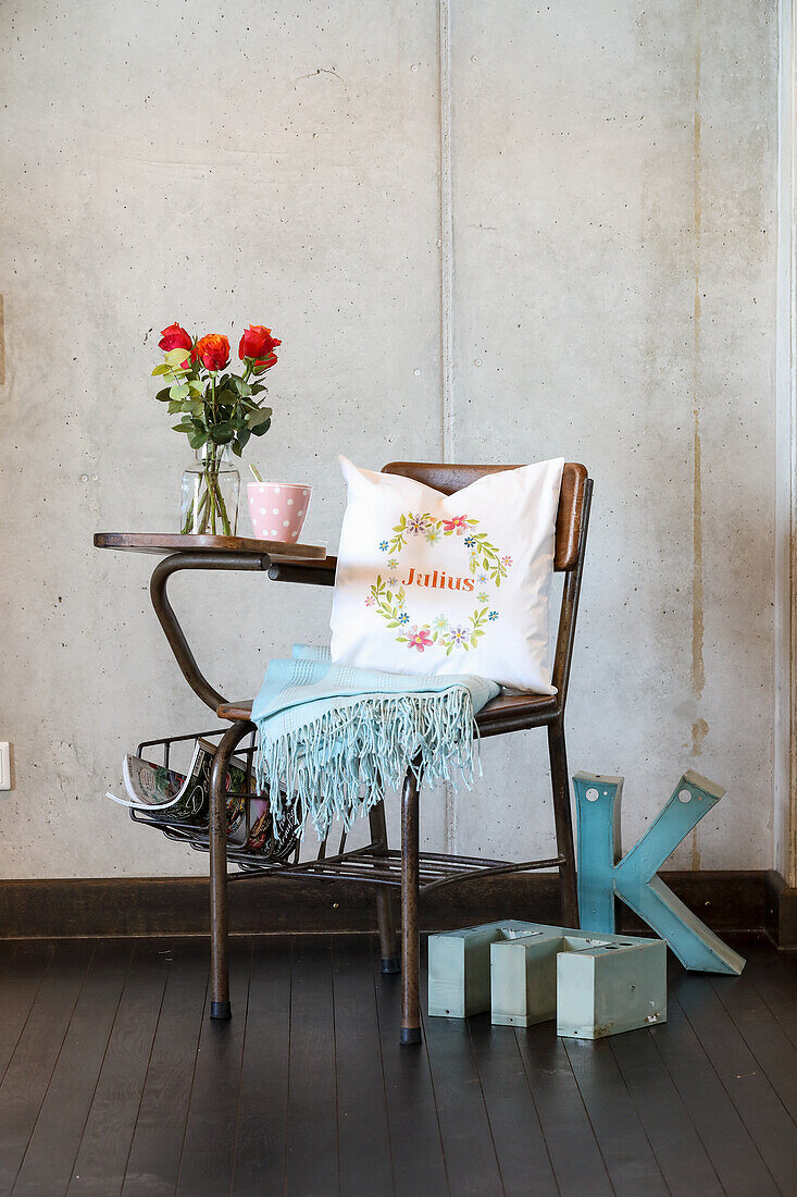 Decorative chair with bouquet of flowers, embroidered cushion and blanket in front of concrete wall