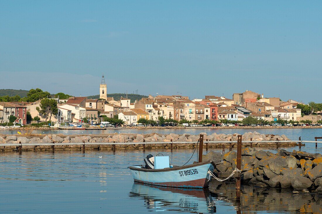 Frankreich, Herault, Bouzigues, auf einem Deich vertäutes Boot mit einem Dorf und seinem Glockenturm im Hintergrund