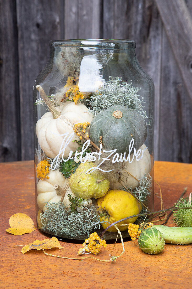 Dekoglas mit Schriftzug "Herbstzauber", gefüllt mit kleinen Kürbissen und Trockenblumen auf Metalltisch