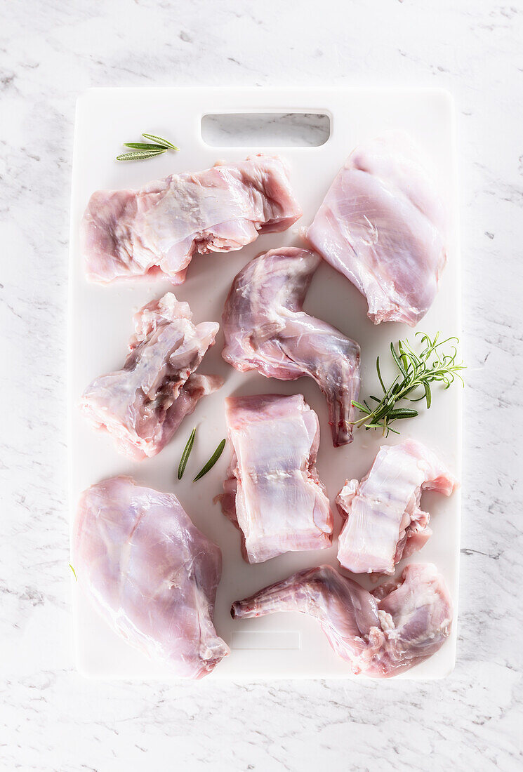 Portioned raw rabbit with rosemary on a white chopping board