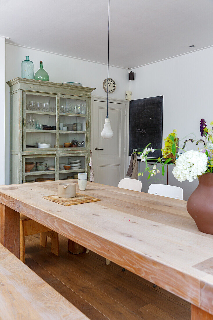 Holztisch mit Blumenvase, Vitrinenschrank und Kreidetafel