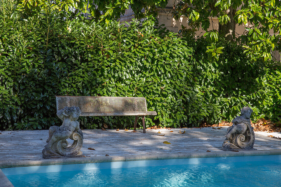 Stone bench and gargoyle statues by the pool in front of an evergreen hedge