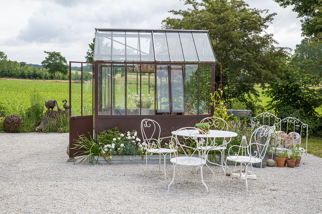 Romantischer Sitzbereich mit weißen Eisenmöbeln vor Gewächshaus im Garten