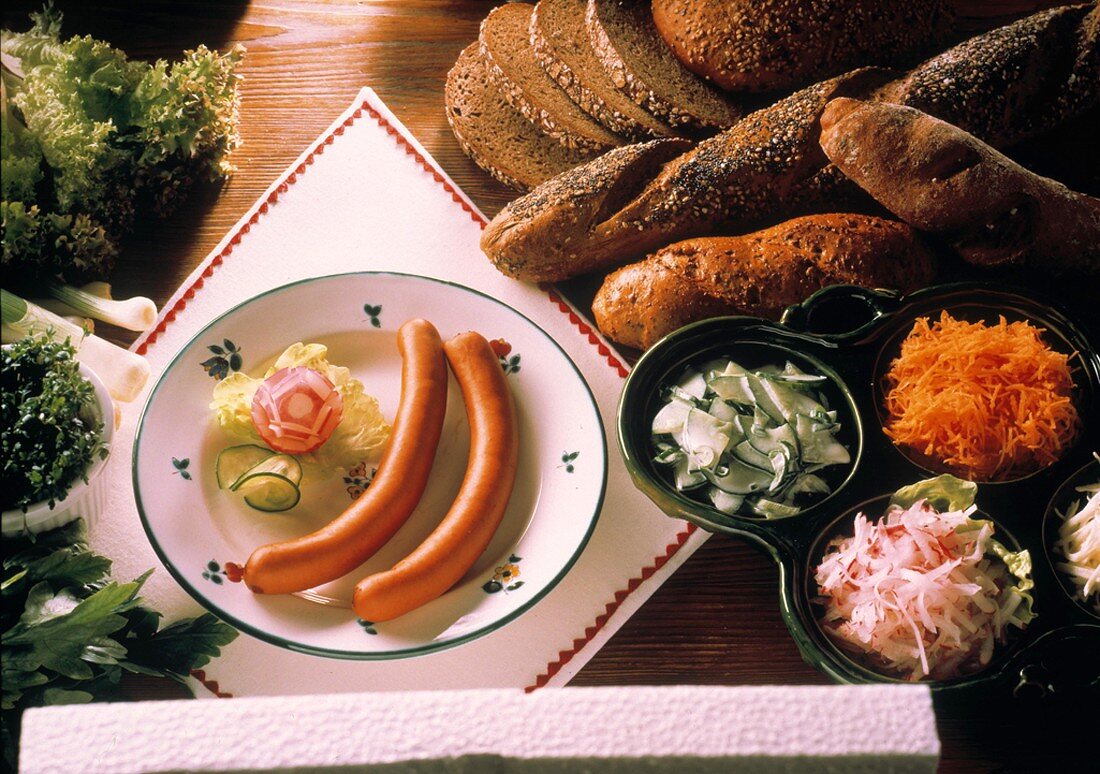 Frankfurter Würstchen auf Teller, daneben Brote, Salate