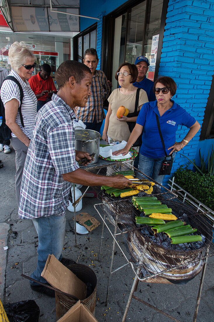 Ein Mann kocht dominikanische Tamales auf einem offenen Grill auf der Straße bei der Bani Mango Expo in Bani, Dominikanische Republik. Eine attraktive blonde ältere Touristin schaut zu