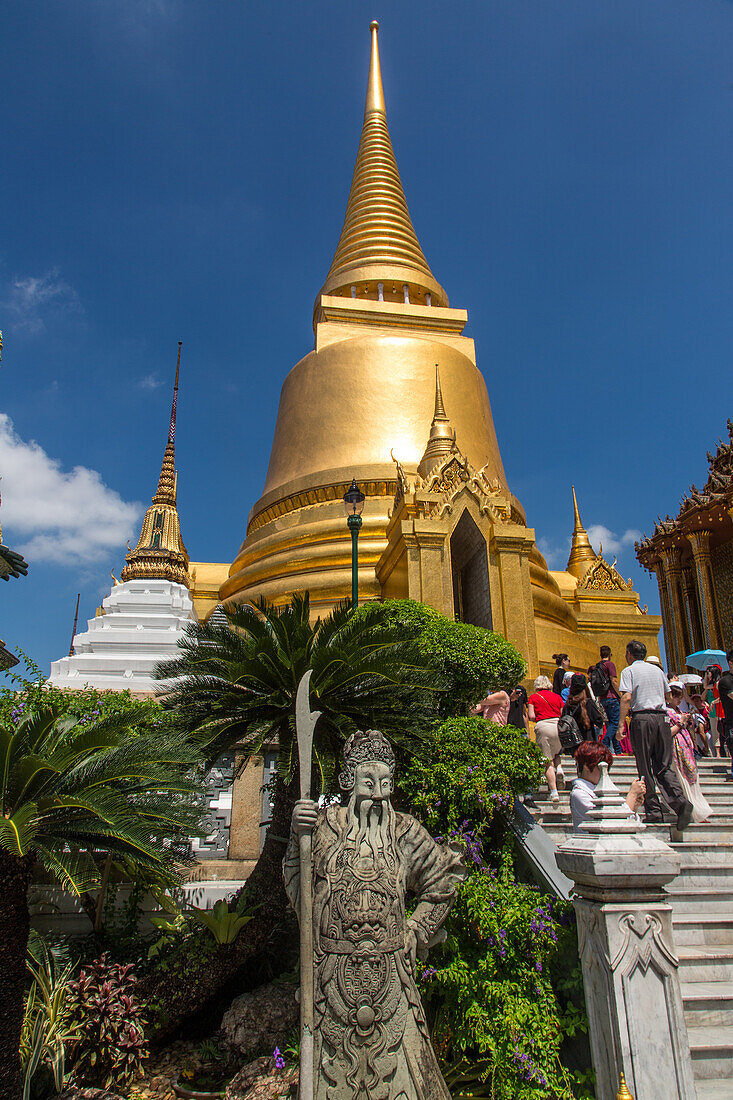 The golden Phra Sri Ratana Chedi by the Temple of the Emerald Buddha at the Grand Palace complex in Bangkok, Thailand. In front is a stone Chinese guardian statue.