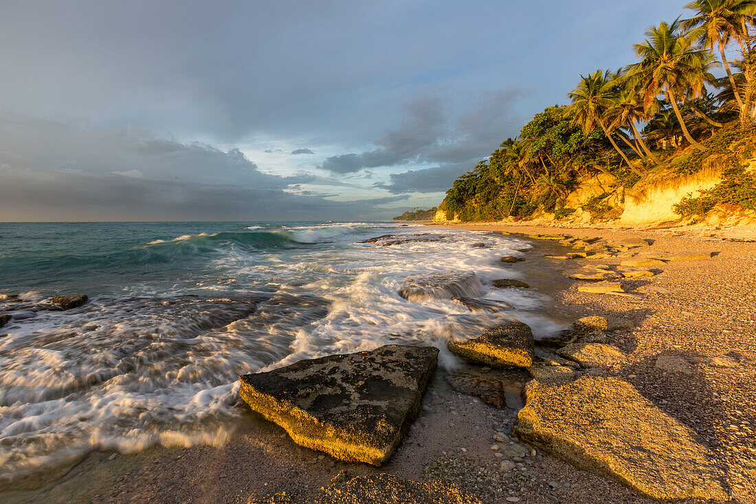 Wellen, die sich bei Sonnenaufgang an einem Strand bei Barahona in der Dominikanischen Republik an den Felsen brechen. Eine lange Verschlusszeit verleiht dem Wasser einen verschwommenen Look