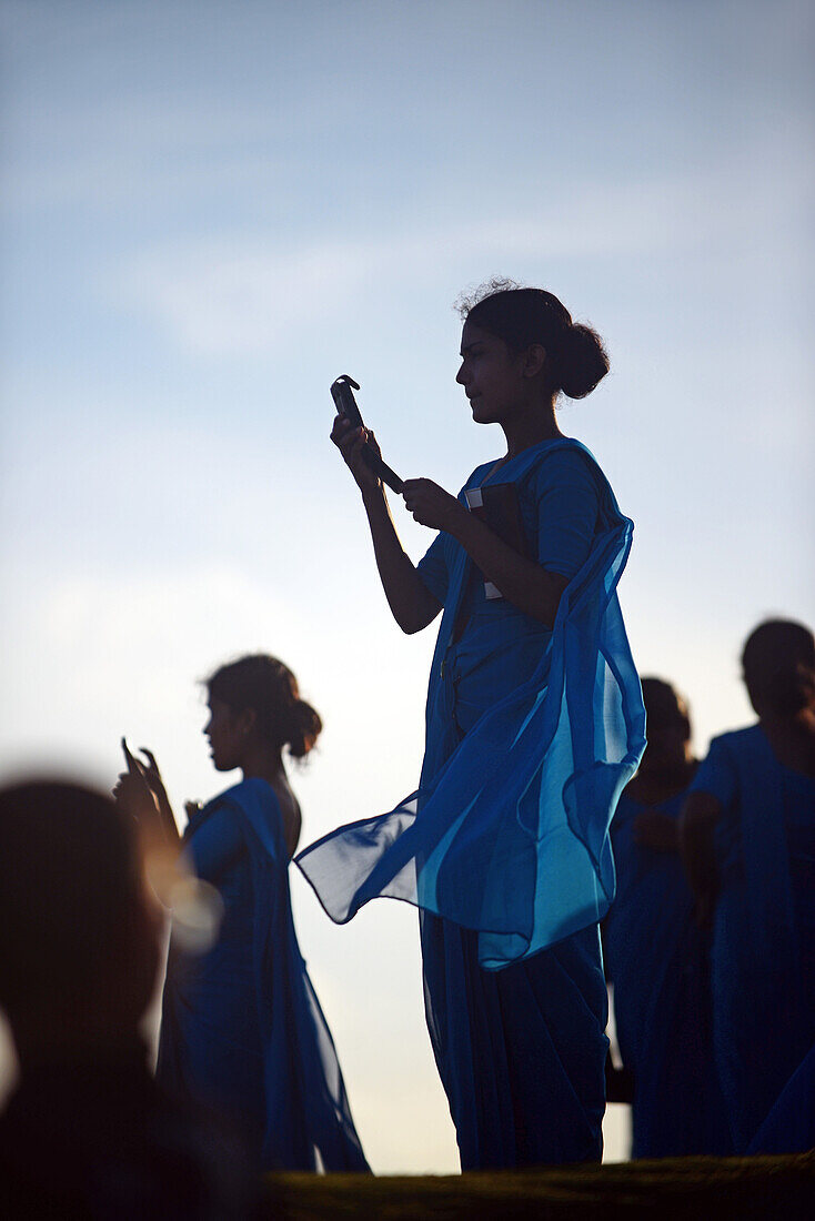 Young woman in numerous group of college students, dressed in blue, uses mobile phone during visit to UNESCO World Heritage, Galle Fort, during Binara Full Moon Poya Day.