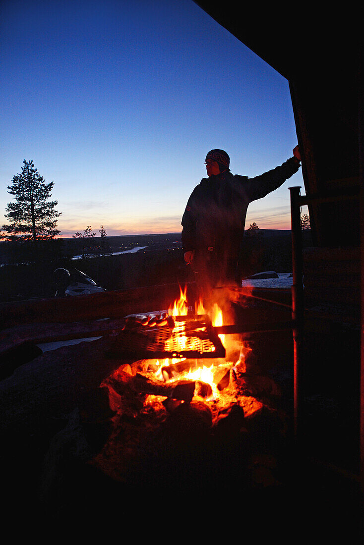 J?nk?l? entspannt sich bei einem Halt am Feuer. Schneemobil-Sonnenuntergangstour mit Arctic Lifestyle, Rovaniemi, Lappland, Finnland