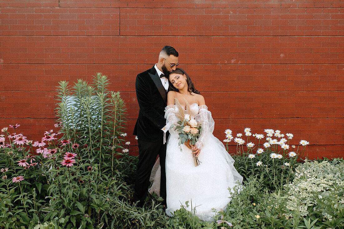 Bräutigam küsst Braut vor einer Mauer und Blumen