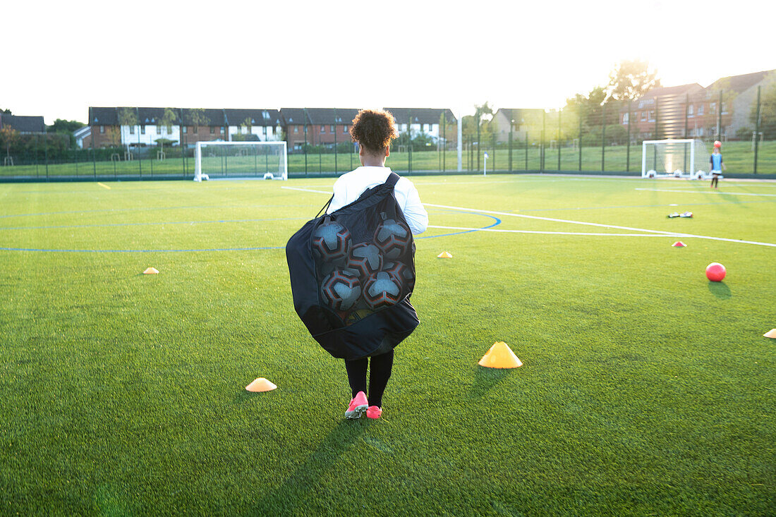 UK, Rückansicht einer Fußballspielerin (12-13), die eine Tasche mit Bällen auf einem Feld trägt