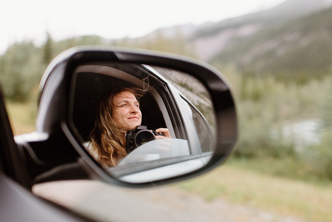 Kanada, Yukon, Whitehorse, Lächelnde Frau mit Kamera im Rückspiegel eines Autos