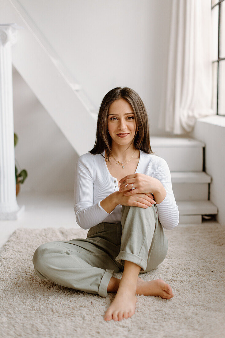 Porträt einer lächelnden jungen Frau, die barfuß auf einem Teppich sitzt