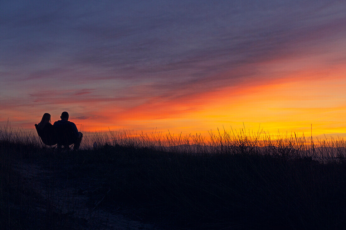 Silhouetten von Mann und Frau, die bei Sonnenuntergang auf einer Bank in einem Feld sitzen