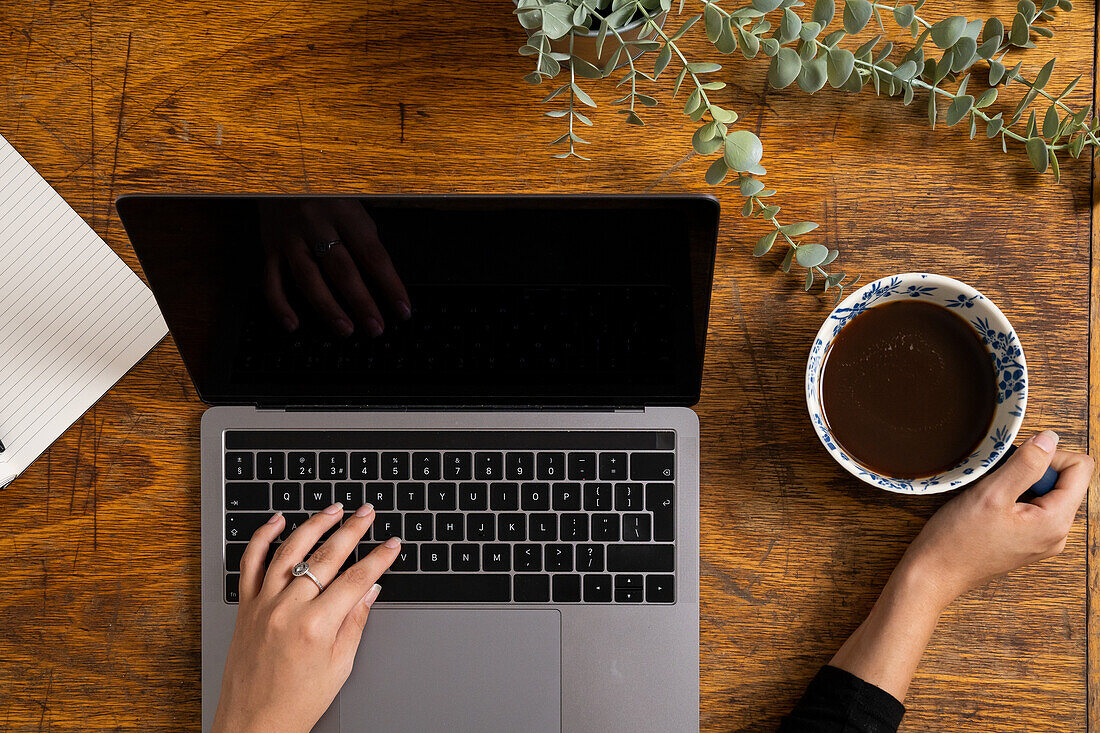 Draufsicht auf die Hände einer Frau, die auf einem Laptop tippt und eine Kaffeetasse hält