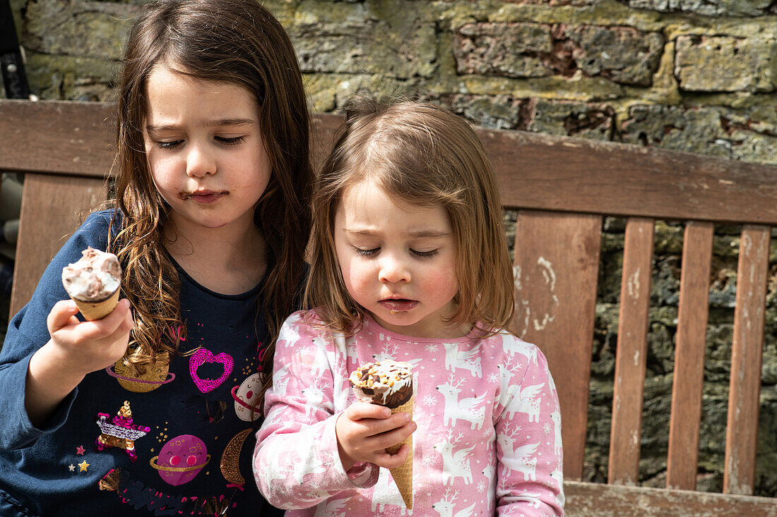 UK, Two girls (2-3, 4-5) with ice cream cones on bench