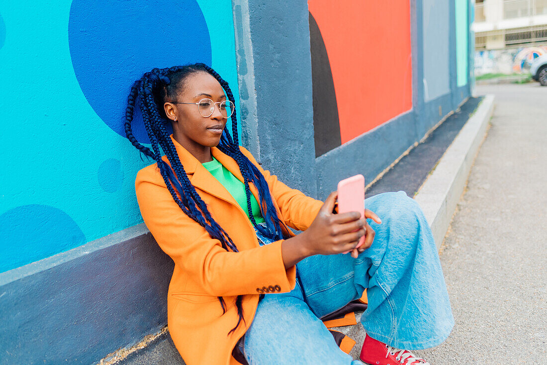 Italy, Milan, Woman with braids sitting by colorful wall, using smart phone