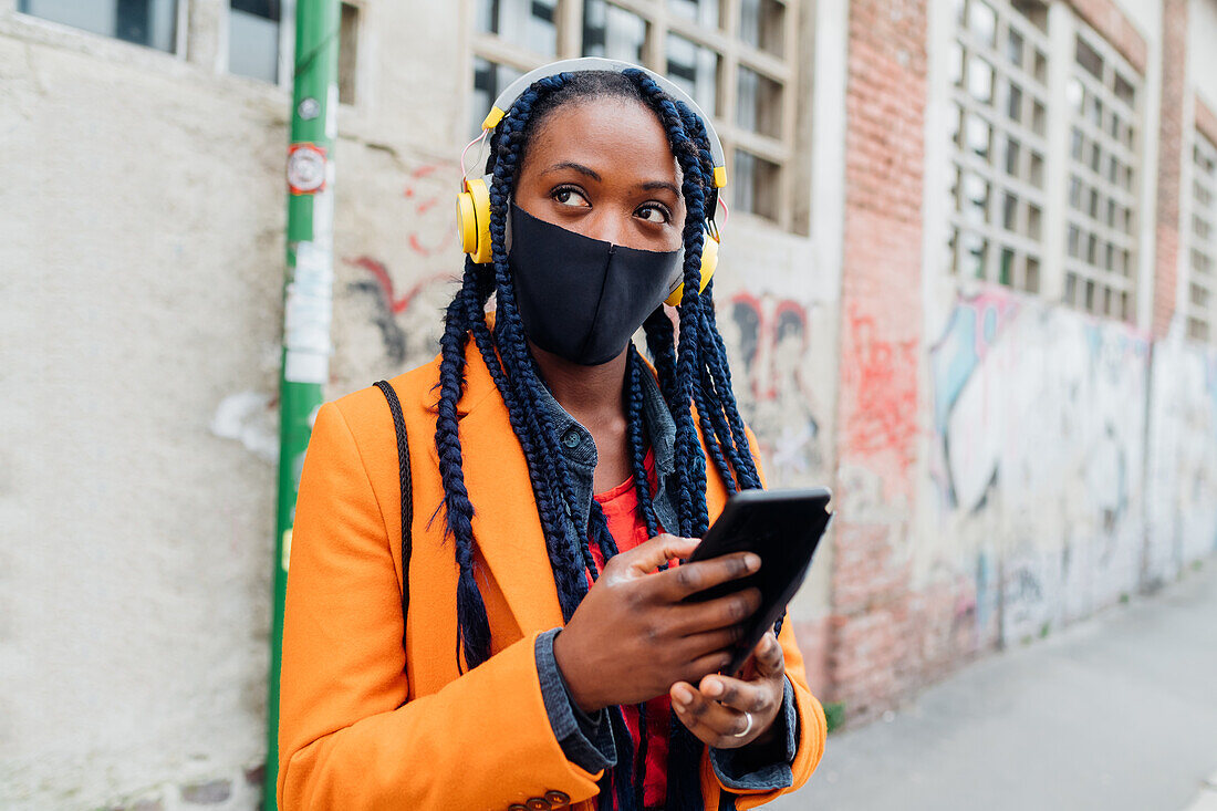 Italien, Mailand, Frau mit Kopfhörern und Gesichtsmaske hält Smartphone