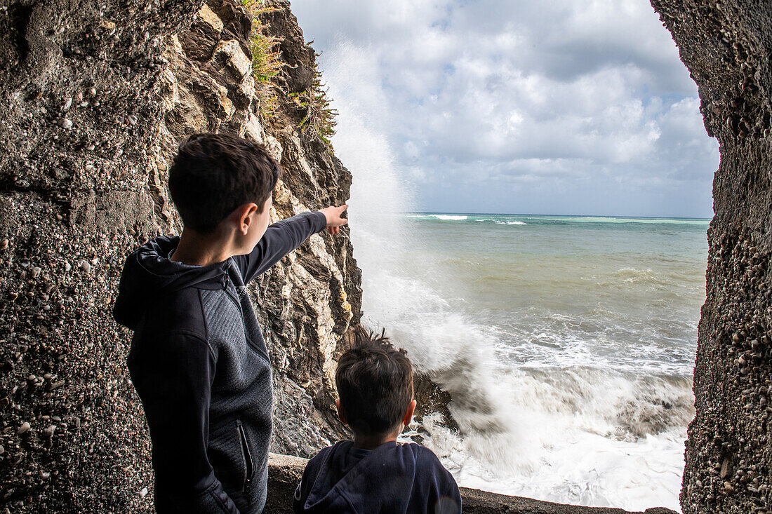 Jungen (6-7, 12-13) schauen auf Meereswellen, die gegen Felsen schlagen, Sizilien, Italien