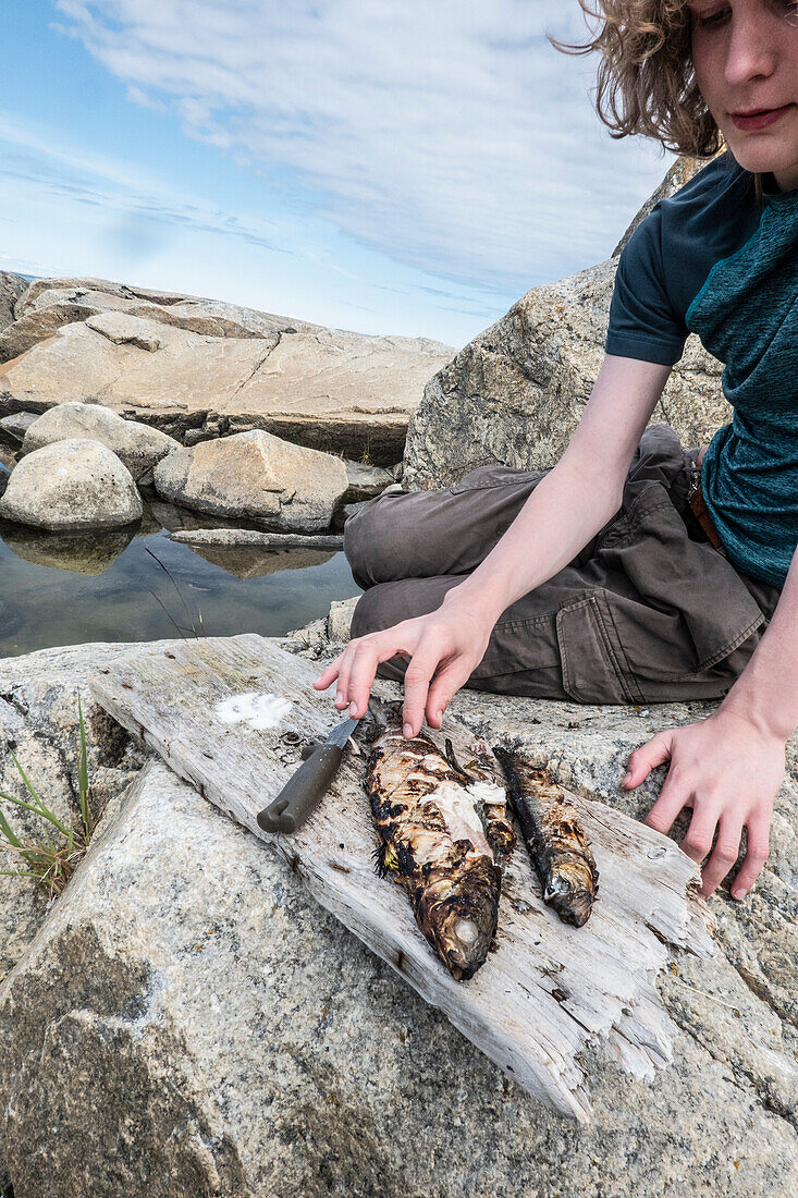 Boy (14-15) sitting on rock and touching grilled fish