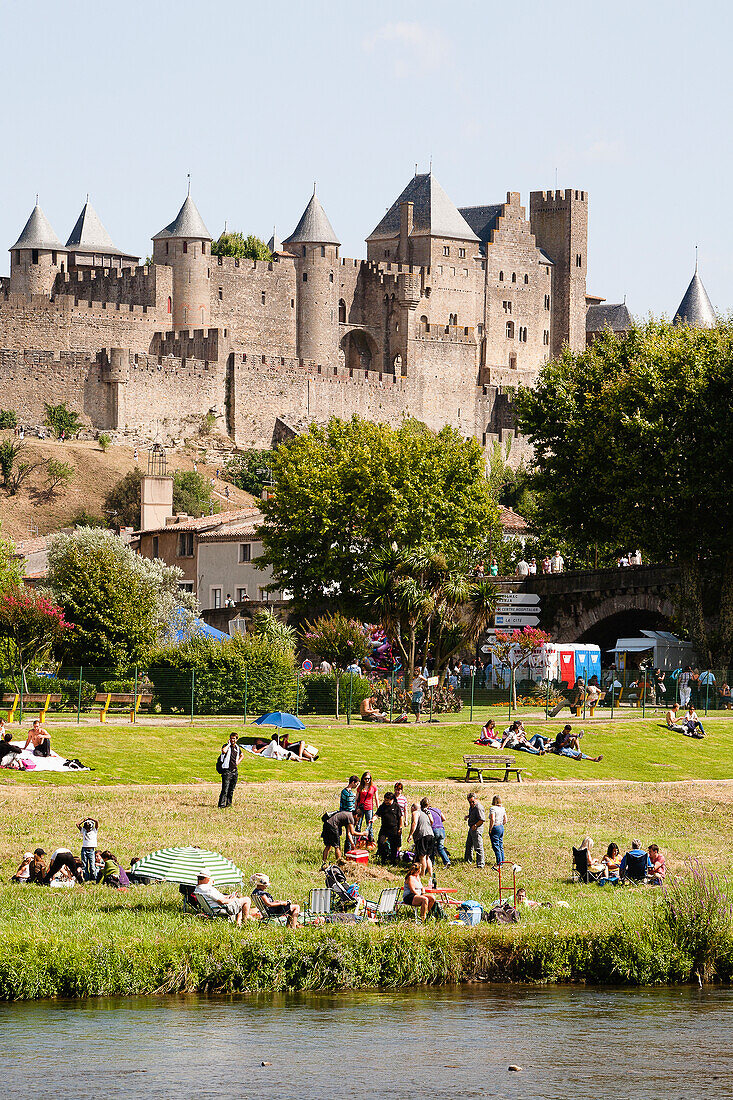 Menschen versammeln sich auf der Wiese am Fluss mit dem Schloss und den Festungsmauern im Hintergrund; Carcassonne, Languedoc-Rousillion, Frankreich