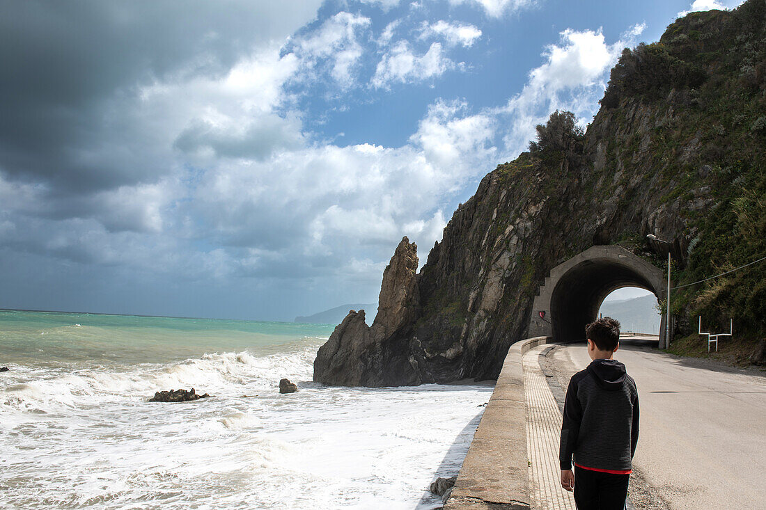 Italien, Sizilien, Junge steht am Straßenrand und schaut auf die Meereswellen