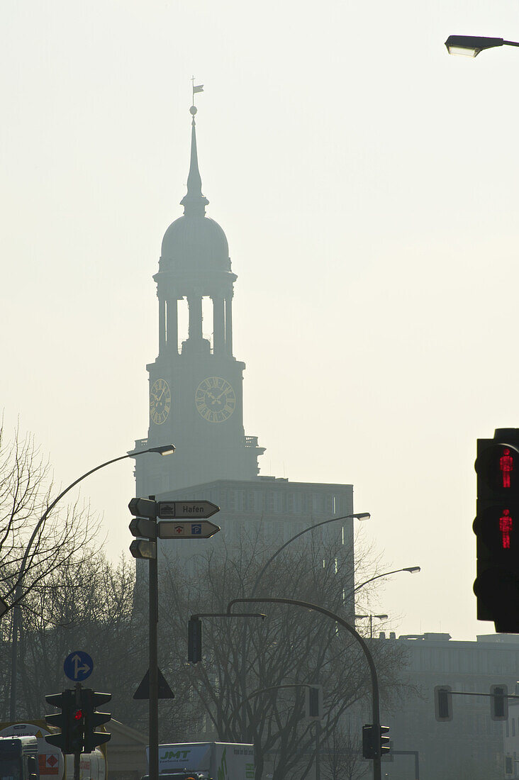 Ein Uhrenturm in der Ferne bei Sonnenaufgang mit Ampeln und Straßenschildern im Vordergrund; Hamburg, Deutschland