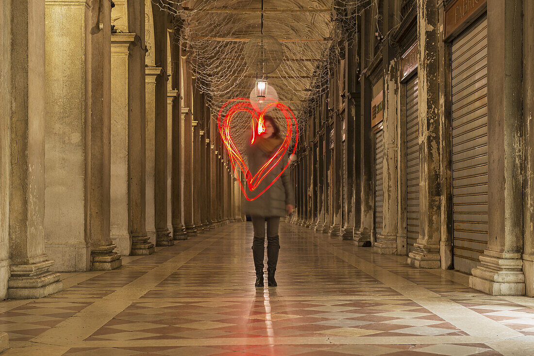 Eine Frau, die in einem Korridor spazieren geht und mit Lichtspuren eine rote Herzform in die Luft zeichnet; Venedig, Venetien, Italien