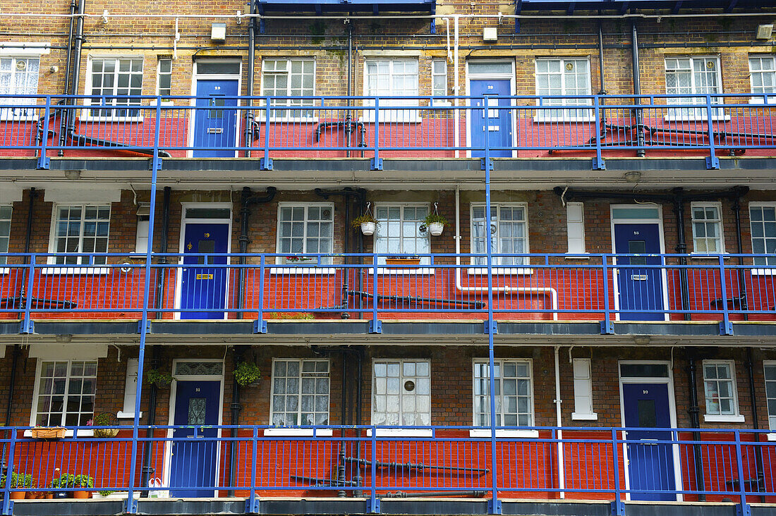 A Brick Residential Building With A Blue Balcony Railings And Doors, And A Red Stripe Painted On The Lower Half Of Each Wall; London, England