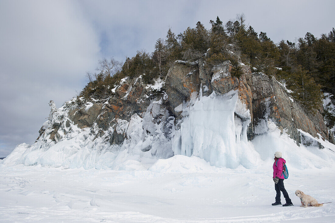 Frau beim Winterwandern auf einem zugefrorenen See in der Nähe eines eisigen Felsufers mit ihrem Hund; Ontario, Kanada