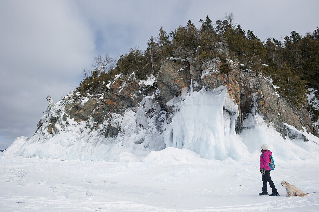 Woman Hiking In Winter On A Frozen Lake Near An Icy Rocky Shore With Her Pet Dog; Ontario, Canada