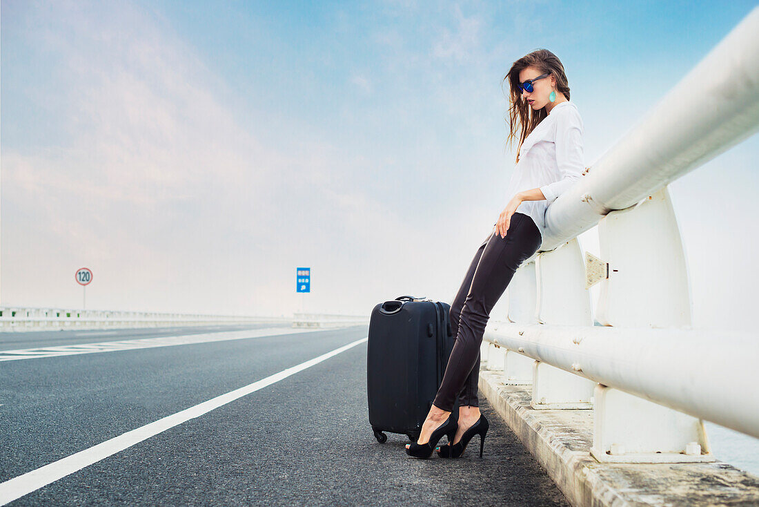 Woman Hitchhiking On A Highway, Resting On A Railing With Her Luggage; Xiamen, Fujian, China